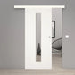A modern white sliding door with a vertical rectangular glass panel, shown installed in a minimalist gray interior doorway with light wood flooring and a black wire chair visible in the background.