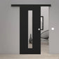 A modern black sliding door with a vertical rectangular glass panel, shown partially open in a minimalist interior setting with light gray walls and light wood flooring.