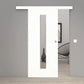 A modern white sliding door with a vertical rectangular glass panel, shown installed in a minimalist gray interior room with light wood flooring.
