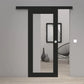 A modern black sliding door with a large vertical glass panel and a small round white doorknob, shown installed in a minimalist gray interior room with light wood flooring.
