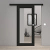 A modern black sliding door with a large vertical glass panel and a small round white doorknob, shown installed in a minimalist gray interior room with light wood flooring.